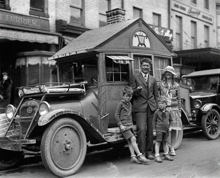 A family of four poses in front of a car converted into a small house, parked on a city street in the 1920s. The father holds a cigar, and the roof sign reads, “Sings and Sells His Own Songs.”