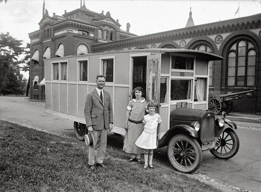 A black-and-white photo of a man and two girls posing in front of an old-fashioned motorhome parked near large brick buildings. The man holds a hat and everyone faces the camera, smiling.