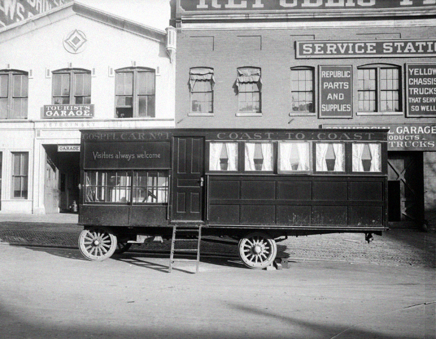 A vintage horse-drawn wagon labeled "Gospel Car" with curtained windows is parked in front of old brick buildings with signs for a service station and garage. A small ladder leads up to the wagon’s door.
