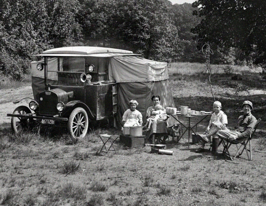 A black-and-white photo of a family camping outdoors beside a vintage car with a tent attachment. The group sits at a table with food, surrounded by camping equipment and trees in the background.