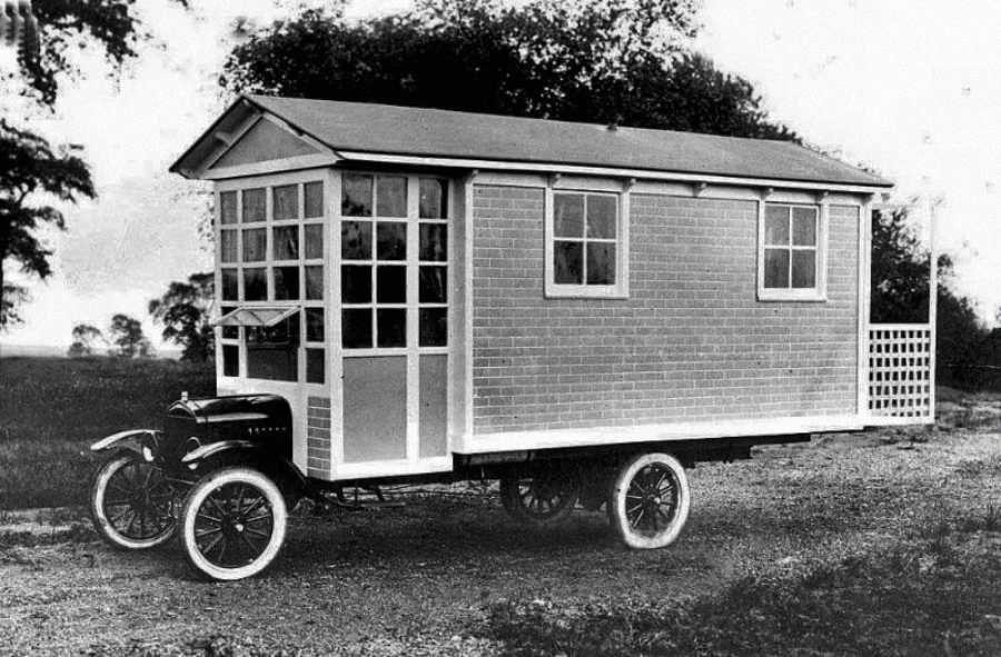 A vintage black-and-white photo shows an old car converted into a house on wheels, resembling a tiny house with windows, a door, and a pitched roof, parked on a dirt road with trees in the background.