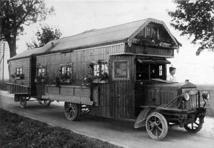 A vintage black-and-white photo of a large, ornate wooden motorhome with flower boxes, resembling a house on wheels. A person is visible driving the vehicle on a rural road lined with trees.