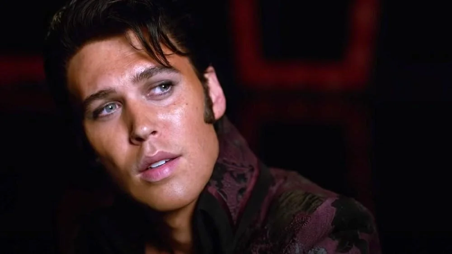 A man with styled dark hair, sideburns, and blue eyes looks to the side. He wears a patterned maroon jacket and has a contemplative expression, set against a dark background.