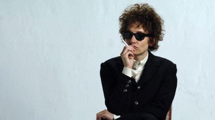 Person with curly hair wearing dark sunglasses and a black suit sits against a plain white background, holding a cigarette close to their mouth with a thoughtful expression.