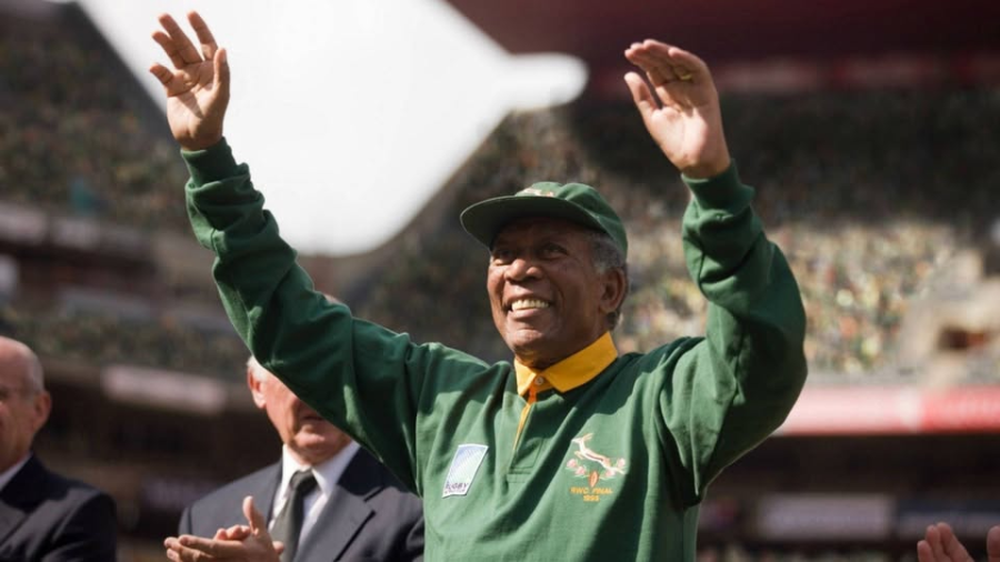 An elderly man in a green sports jacket and cap smiles and raises both hands in celebration at a stadium, with people and a crowd blurred in the background.
