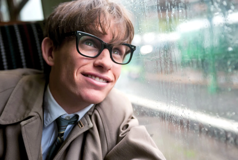 A young man with tousled hair, wearing large black glasses and a beige coat, smiles while looking out a rain-streaked train window.