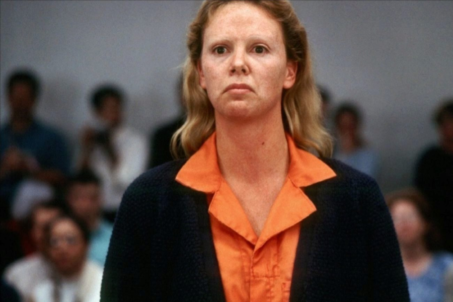 A woman with light hair, wearing an orange shirt and a dark jacket, stands in a courtroom with a serious expression. Blurred people are seated in the background.