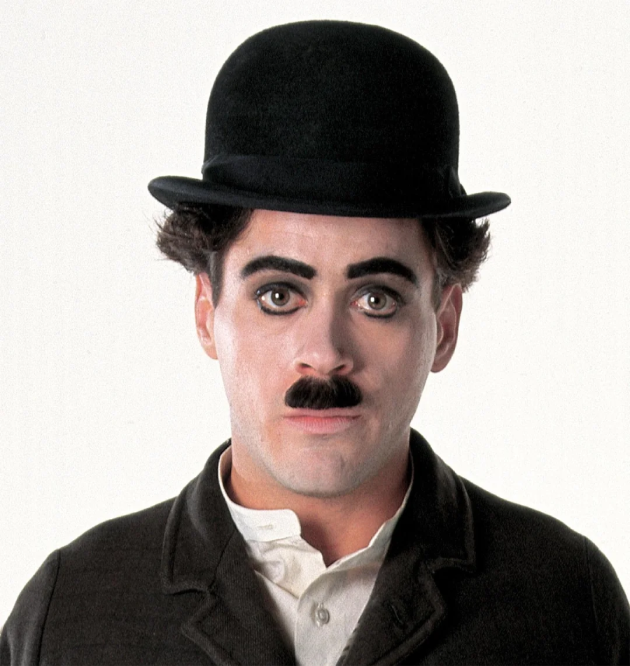 A person dressed as Charlie Chaplin's iconic character, wearing a black bowler hat, black coat, white shirt, black mustache, and exaggerated eyebrow makeup, stands against a plain white background.