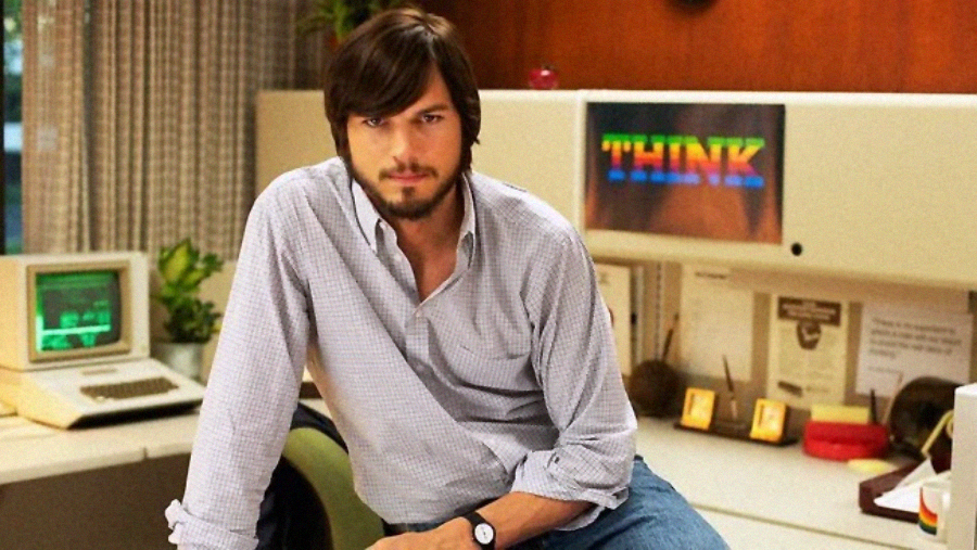 A man with dark hair and a beard sits on a desk in an office with vintage computers. Behind him, a monitor displays the word "THINK" in rainbow colors. The setting appears retro, with wood paneling and old technology.