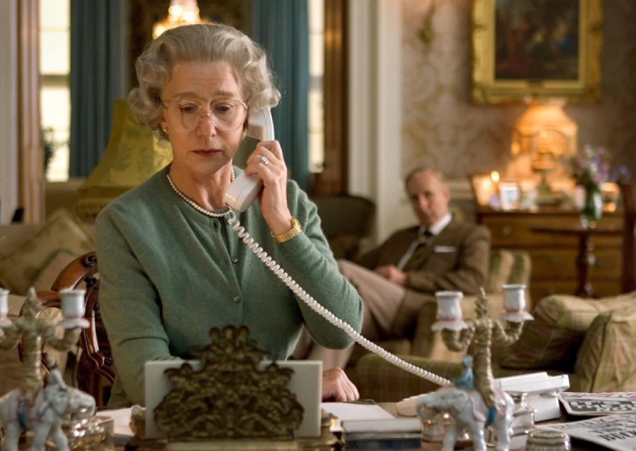 An older woman with short gray hair and glasses, dressed formally, speaks on a corded phone at a desk cluttered with papers and figurines. A man sits in the background on a couch in a warmly lit, elegant room.