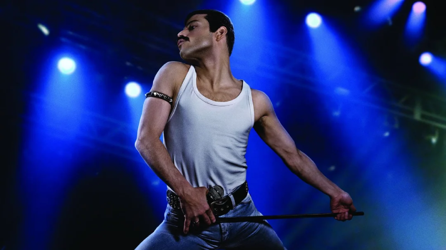 A person in a white tank top, jeans, and armband strikes a dramatic pose on stage under bright blue stage lights, holding a microphone stand.