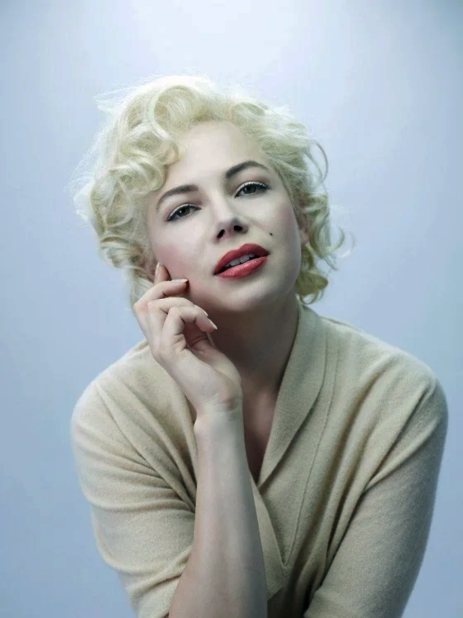 A woman with short, curly platinum blonde hair and red lipstick gazes softly at the camera, resting her chin on her hand. She wears a beige top against a light blue background.