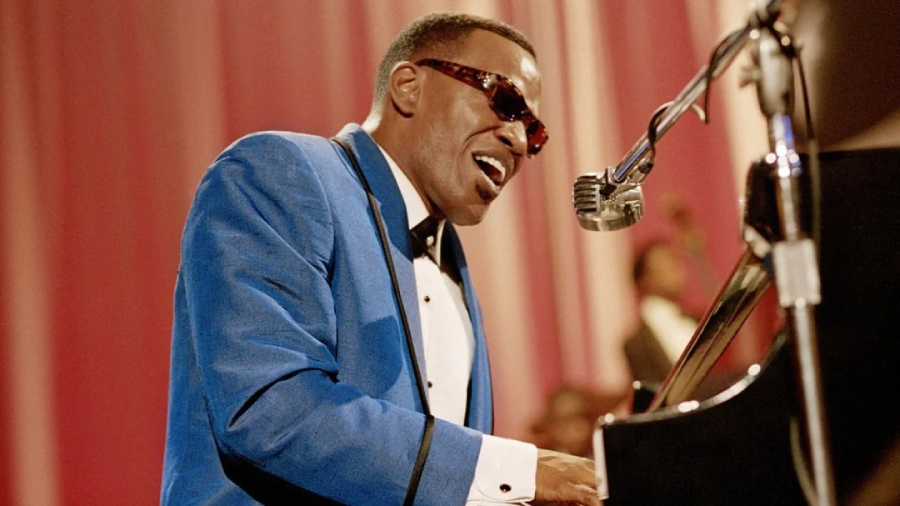 A man in a blue suit and sunglasses plays the piano and sings passionately into a vintage microphone, with pink and white curtains in the background.