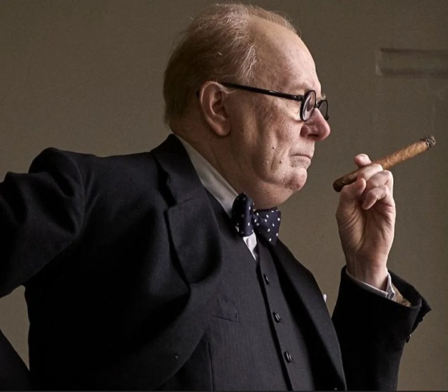 A man wearing glasses, a dark suit, and a polka dot bow tie stands in profile, holding a cigar near his face, looking thoughtful.