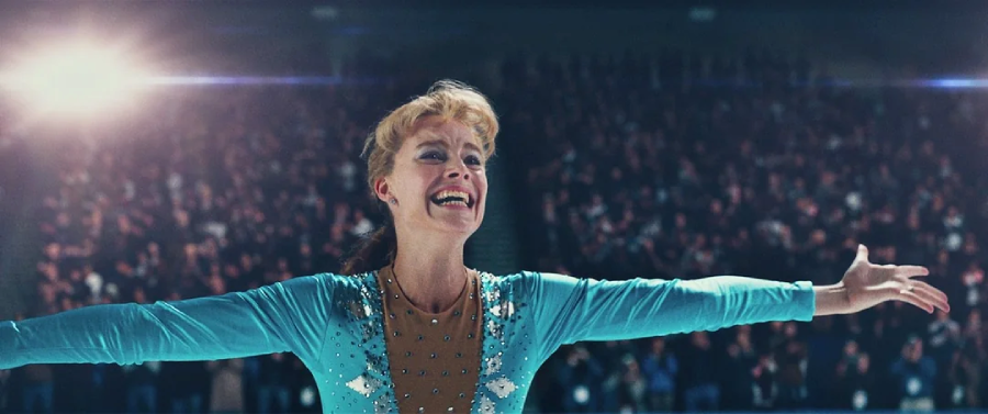 A female figure skater in a blue costume stands on the ice with arms outstretched, smiling broadly. The background shows a cheering audience in a dimly lit arena.