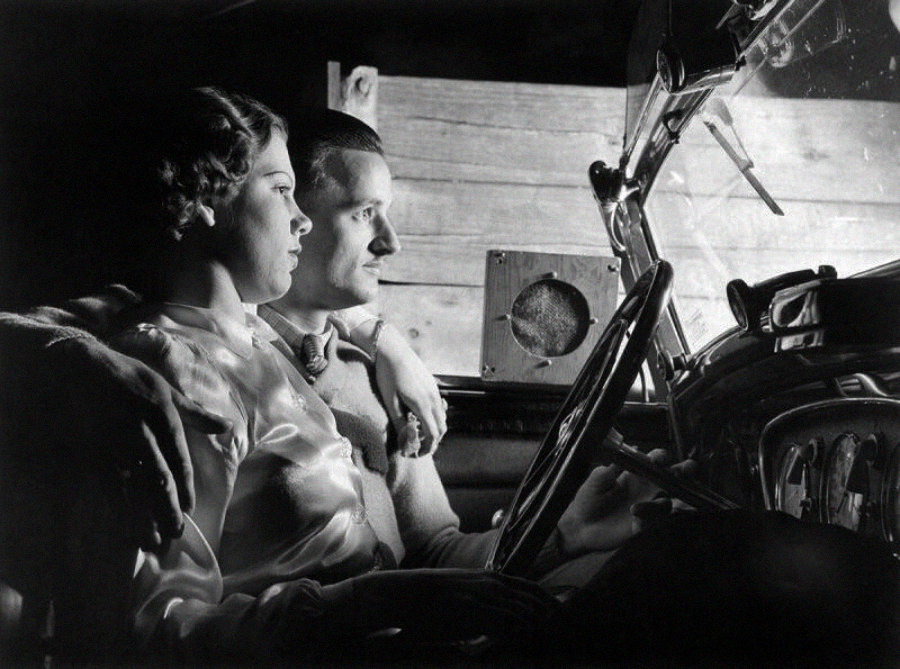 A black-and-white photo of a woman and a man sitting close together in the front seat of a car at night, illuminated by light from outside the vehicle. The man has his arm around the woman as they look forward.