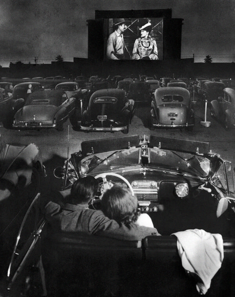 Black and white photo of people sitting in classic cars at a drive-in movie theater at night, watching a film projected on a large outdoor screen. The image is viewed from behind a convertible.