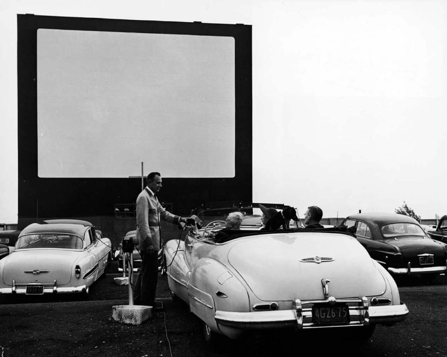 A black-and-white photo of a man taking payment from people in a convertible car at a drive-in movie theater, with other vintage cars parked in front of a large blank movie screen.