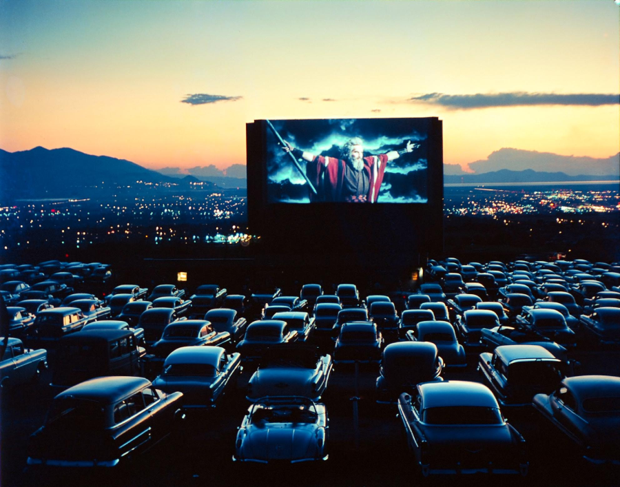 A drive-in theater at dusk with rows of vintage cars facing a large movie screen showing a dramatic scene of a man holding a staff, as city lights glow in the background beneath a colorful sky.
