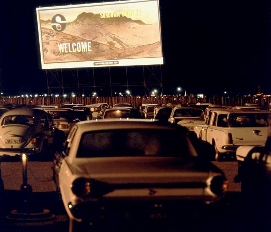 Rows of classic cars parked at a drive-in theater at night, facing a large illuminated screen displaying a "Welcome" message and a mountain landscape.