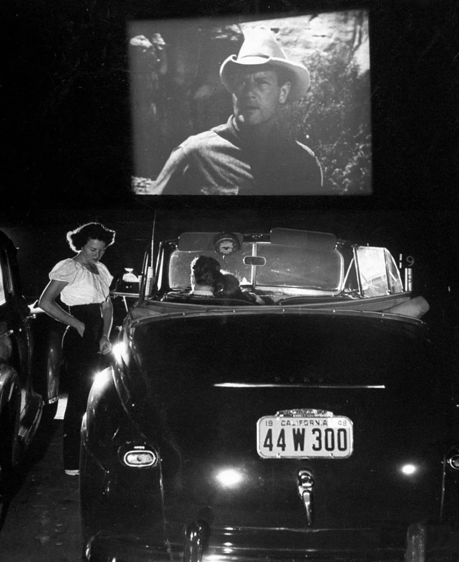 A woman stands beside a convertible car at a drive-in movie theater at night, with a black and white film featuring a cowboy projected on a large outdoor screen in the background.