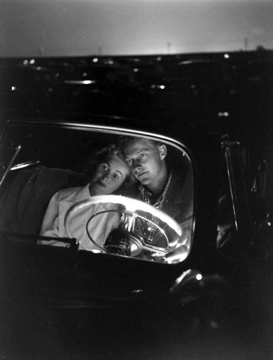 A black-and-white photo of a young couple sitting close together in a car at night, illuminated by the steering wheel and dashboard lights, with a dark parking lot visible through the windows.