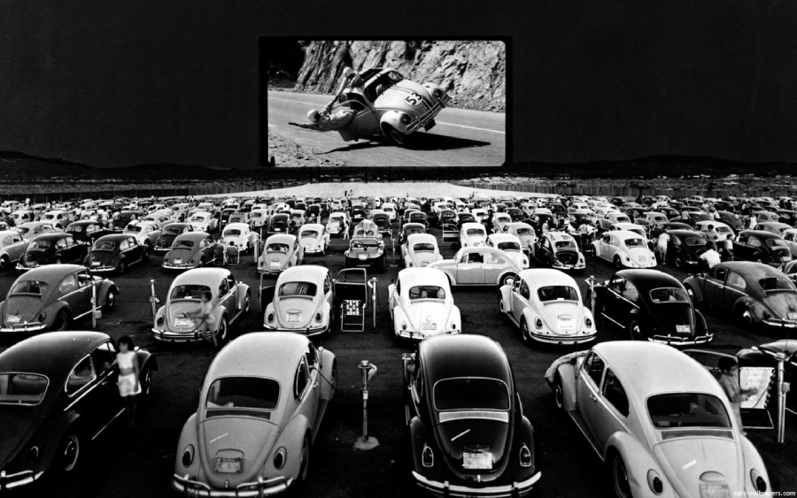 A crowded drive-in theater at night shows a movie featuring cars on a large screen. Rows of classic cars, mostly Volkswagen Beetles, are parked as people watch the film outdoors.