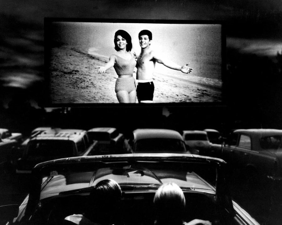 A black-and-white photo of a drive-in theater shows cars parked facing a large outdoor screen, which displays a smiling man and woman in swimwear posing on a beach. Two people watch from a convertible in the foreground.