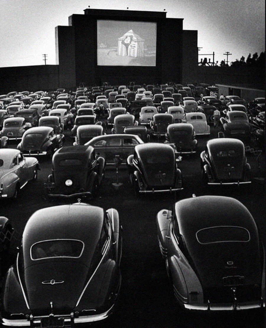 Black and white photo of a crowded drive-in theater at night, with many vintage cars parked and people watching a movie projected on a large outdoor screen in the distance.