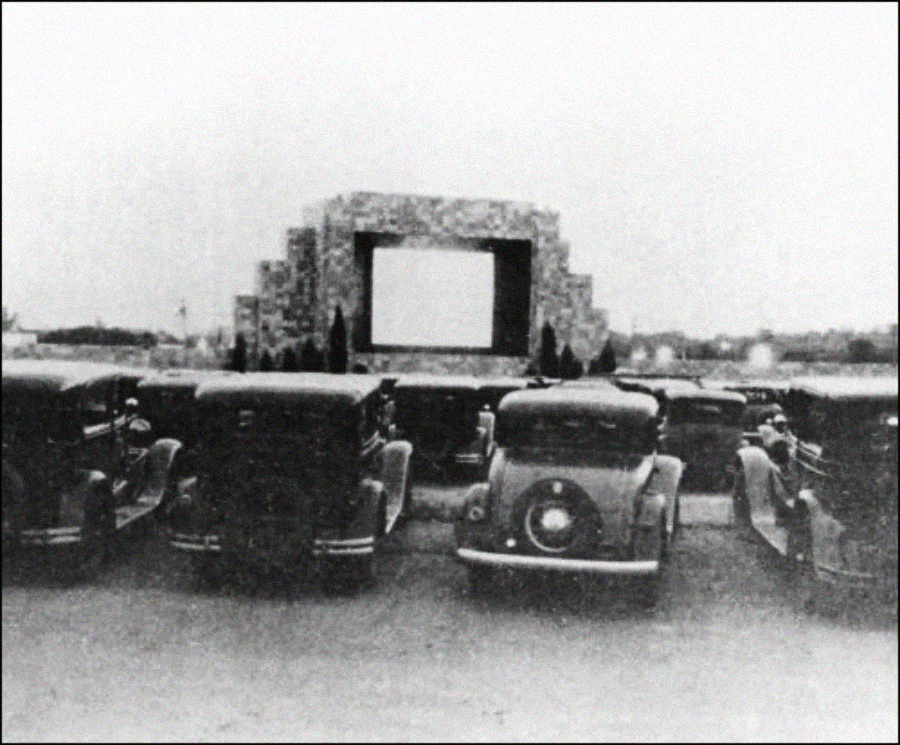 A black-and-white photo of vintage cars parked in front of a large outdoor movie screen at a drive-in theater, with trees and open sky in the background.