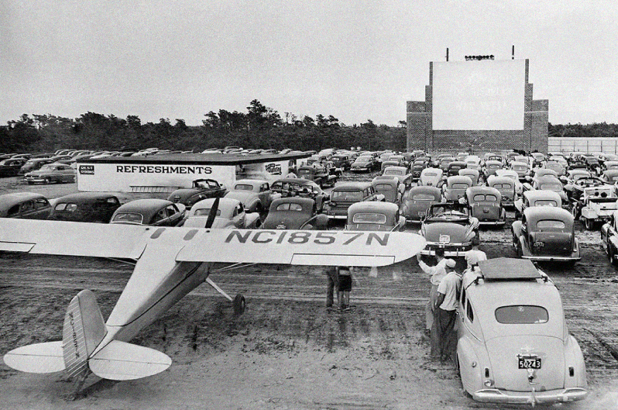 Black-and-white photo of a crowded drive-in theater parking lot, showing vintage cars and a small airplane parked among them. People stand near the airplane and a nearby refreshment stand. The movie screen is visible in the background.