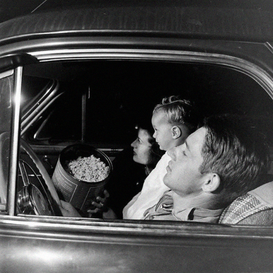 A man, woman, and young boy sit in a parked car at night, sharing a large tub of popcorn. The man is in the driver’s seat, the woman and boy look out the passenger window. They appear to be at a drive-in movie.