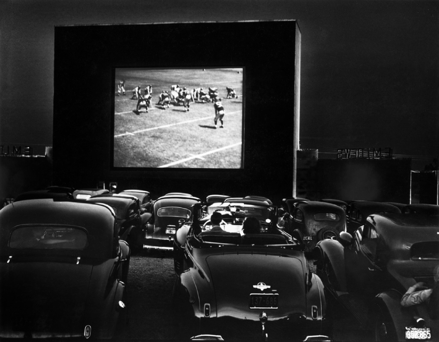 Black-and-white photo of cars parked at a drive-in theater at night, with people watching a film featuring a football game projected on a large outdoor screen.