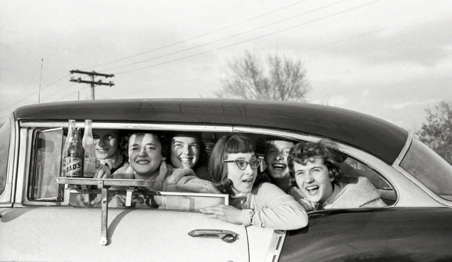 A group of smiling young women lean out of a car window, laughing and posing together. Two glass soda bottles are placed on a tray attached to the car door. The scene appears to be from the mid-20th century.