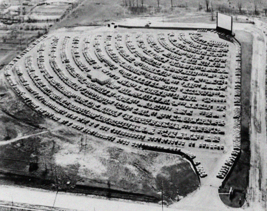 A black-and-white aerial view of a large drive-in movie theater shows hundreds of cars parked in curved rows facing a large outdoor screen, surrounded by open land.