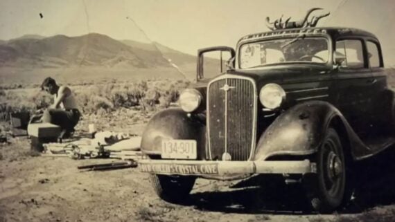 A black and white photo shows an old car with a visible front license plate and deer antlers on the hood, parked on a dirt road with mountains in the background. A person is sitting on the ground, working with equipment.