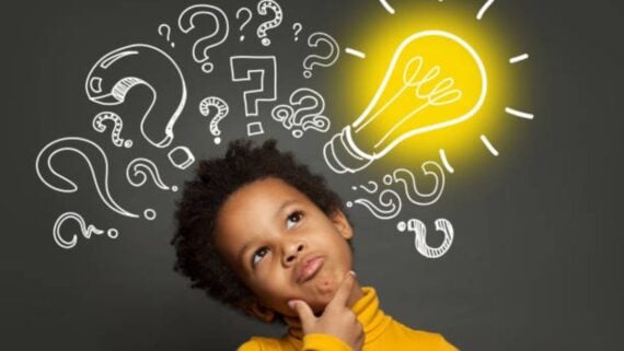 A child in a yellow shirt looks thoughtful, with hand on chin, in front of a dark background with chalk-drawn question marks and a large, glowing light bulb above their head.