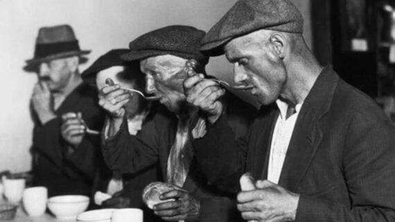 Four men in hats and worn clothing sit side by side, eating soup with spoons from bowls. They appear focused on their meal. The image is black and white and has a historical, vintage feel.