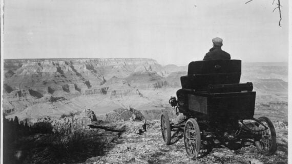 A person sits in an early vintage car parked on the edge of the Grand Canyon, looking out over the vast canyon landscape under a clear sky.