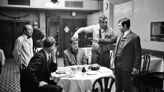 Five men are in a restaurant; one is sitting at a table with a gun pointed at his head by another man while a third holds his hair. Two other men watch, one covering his face. The scene is tense and dramatic.