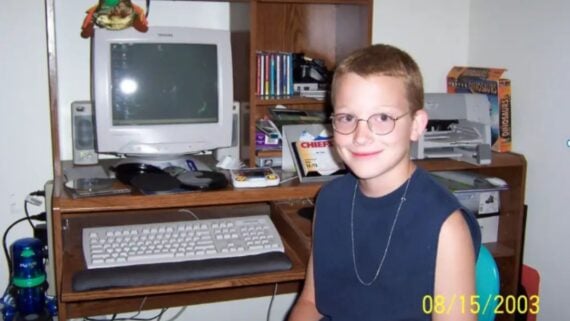 A young boy with glasses and a sleeveless shirt smiles while sitting at a desk with a vintage computer, keyboard, and various office supplies. The photo is dated August 15, 2003.