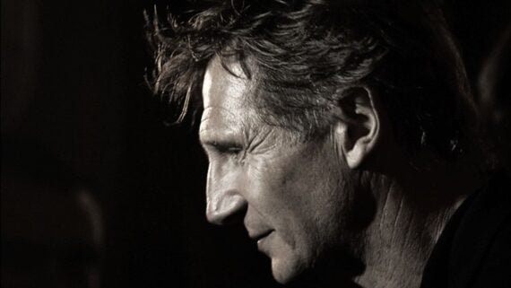 A grayscale close-up of a middle-aged man with tousled hair, shown in profile against a dark background, looking downward with a thoughtful expression.