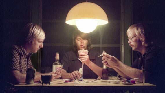 Three young men sit around a table playing cards under a round hanging lamp. Bottles, poker chips, and drinks are on the table. One man holds cards, another rolls a cigarette, and the third studies his hand.