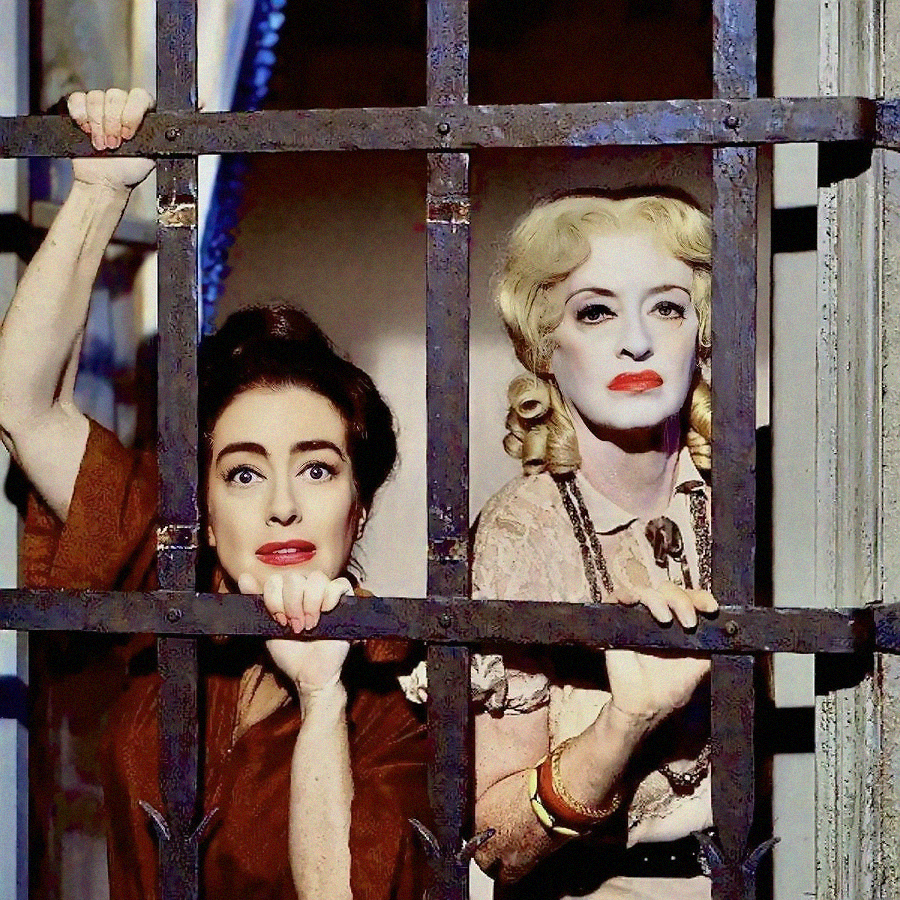 Two women with dramatic makeup and expressions stand behind metal bars, gripping them with their hands. One has dark hair and the other has blonde hair styled in curls. Both look out through the bars with intense, worried gazes.