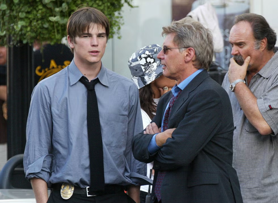 Two men stand talking outdoors; one wears a blue shirt, tie, and police badge on his belt, while the other wears a suit and sunglasses. A woman in a floral hat and a man with a beard are in the background.