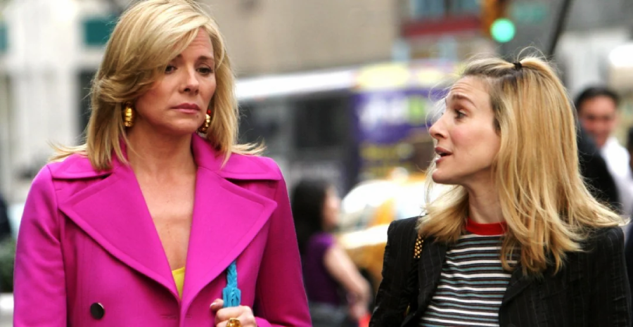 Two women walk side by side on a city street. One wears a bright pink coat and gold earrings, while the other has long blonde hair and wears a striped top. They appear to be engaged in conversation.