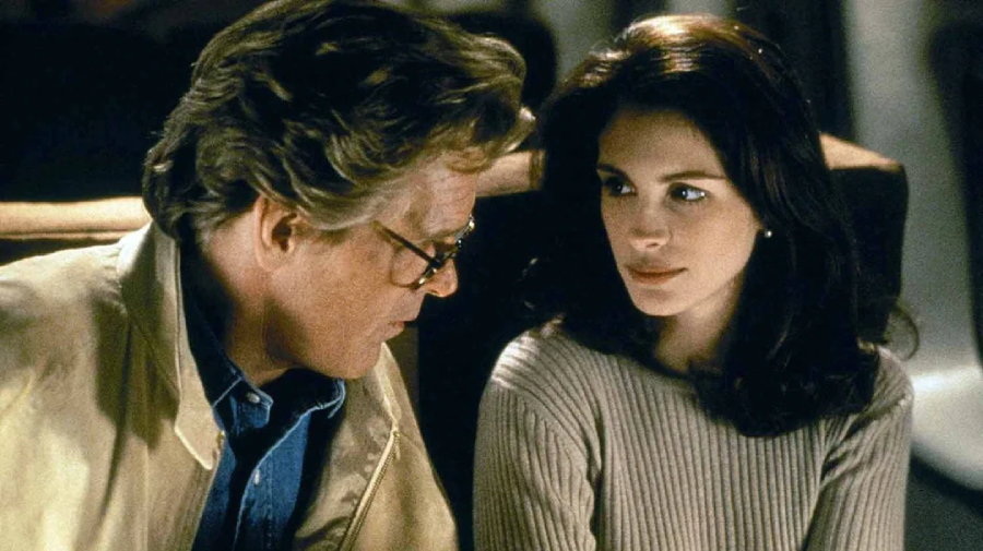 A man with glasses and a light jacket sits closely next to a woman with dark hair and a beige sweater. They are indoors, and the woman looks at the man attentively as he speaks.