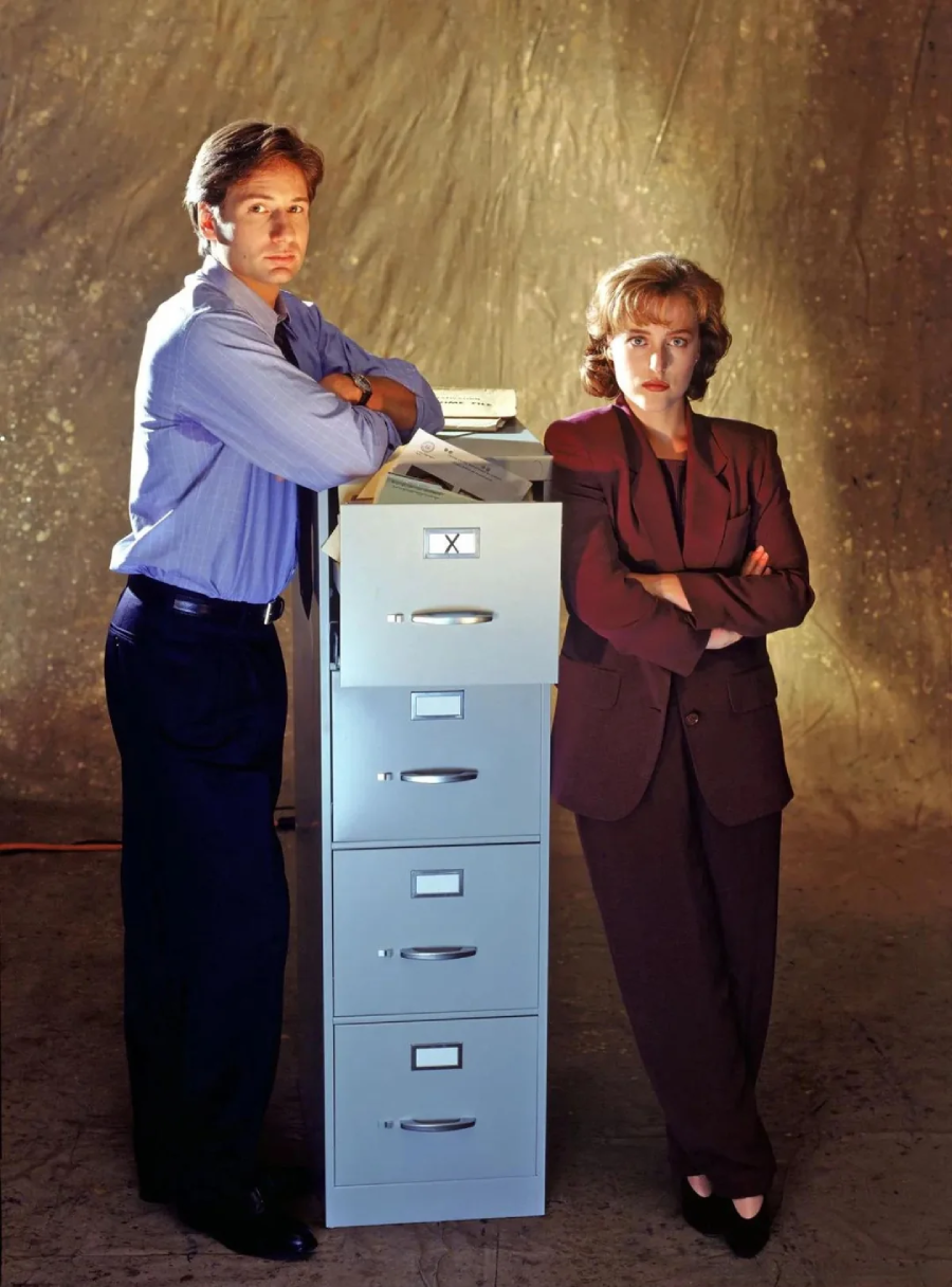 A man in a blue shirt and tie and a woman in a brown suit stand with arms crossed next to a tall filing cabinet against a brownish backdrop, both looking directly at the camera.