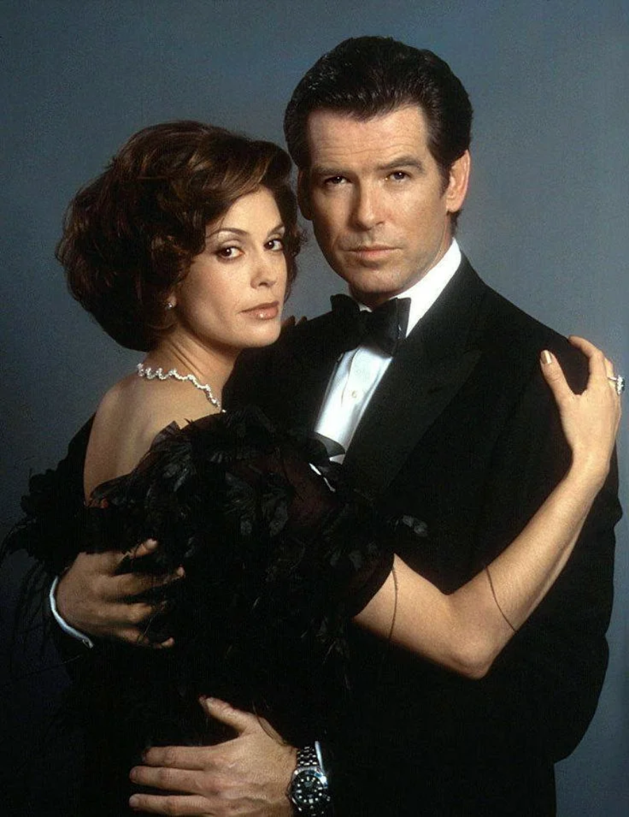 A man in a black tuxedo and a woman in a black feathered dress stand close together, posing against a grey background. Both have serious expressions and are elegantly dressed.