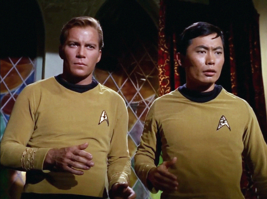 Two men wearing gold Starfleet uniforms stand side by side in a room with stained glass windows and red curtains, looking intently ahead.